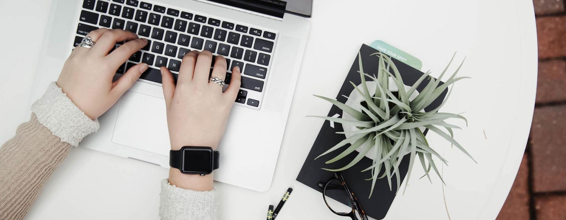 overhead shot of woman working on a laptop at a round table with pencils, her cell phone, glasses, notebook and a potted plant