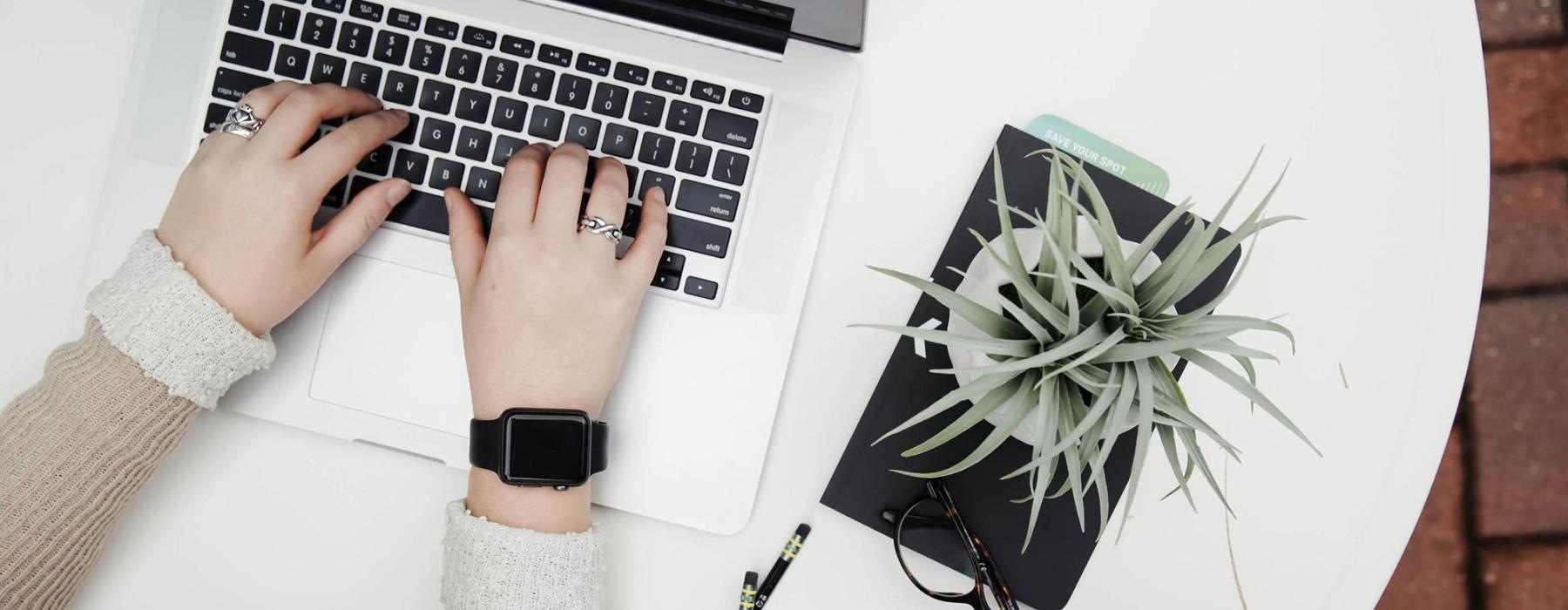 overhead shot of woman working on a laptop at a round table with pencils, her cell phone, glasses, notebook and a potted plant